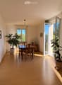 Bright dining area with wooden floor, large windows, dining table with chairs, potted plants, and a cabinet.