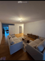 Modern living room with light-colored wooden flooring, a large sectional sofa, a coffee table, and a side table with decorative items. The room features white walls and a ceiling light fixture.
