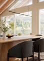 Modern dining area with a large wooden table, grey upholstered chairs, a vase with greenery, and a bowl of fruit. The space features exposed wooden beams, large windows with a scenic mountain view, and contemporary kitchen elements.