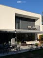 Modern house exterior with a spacious patio area featuring a dining table, chairs, a sofa, and potted plants. The house has large windows and a balcony with additional seating.