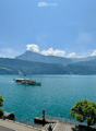 A scenic view of a lake with a boat sailing on it, surrounded by lush green trees and distant mountains under a clear blue sky.
