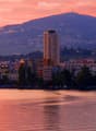 A scenic view of a cityscape at sunset, featuring a body of water in the foreground, with buildings and trees lining the shore, and mountains in the background.