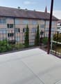 Balcony with metal railing, concrete floor, overlooking a residential area with buildings and trees.