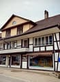 Two-story building with a tiled roof, multiple windows, and a storefront with awnings and signage. The ground floor has glass windows displaying various items, and the upper floor features balconies and decorative railings.