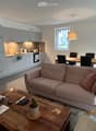 A modern, open-concept living and dining area with a light pink sofa, wooden coffee table, dining table with chairs, and a small kitchen area with white cabinets and stainless steel appliances.