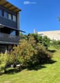 Lush green garden with well-maintained lawn, various shrubs, and trees, featuring a modern multi-story building with balconies and large windows in the background.