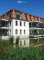 Modern two-story building with white walls, red-tiled roof, and large glass balconies overlooking a serene water body.
