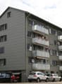Four-story apartment building with grey facade, multiple balconies, and flower boxes. Several cars and a motorcycle are parked in front of the building.