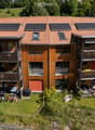 Modern multi-story building with wooden balconies, solar panels on the roof, and a landscaped garden area.