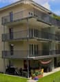 Modern three-story apartment building with multiple balconies, surrounded by greenery and a small garden area with outdoor furniture.