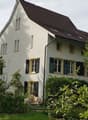 Two-story white house with a brown tiled roof, multiple windows with shutters, and a well-maintained garden in front.