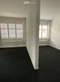 Empty room with white walls, dark carpet, large windows with blinds, and a radiator under one window.