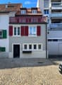 Two-story house with light gray walls, red shutters, and a red balcony railing. The ground floor has a black door and several windows, while the upper floor features additional windows and a small balcony. The roof is tiled with brown tiles and has dormer