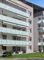 Modern multi-story apartment building with glass balconies, light-colored facade, and surrounding green lawn.