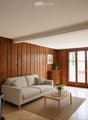 Modern living room with wooden walls and floor, large sliding glass doors leading to a balcony, a light-colored sofa, a wooden coffee table, and decorative plants.