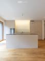 Modern, minimalist kitchen with white cabinetry, stainless steel appliances, and a central island. Large windows provide natural light, and the floor is made of light wood.