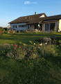 A well-maintained garden with a lawn, flower beds, and a house in the background. The house has a tiled roof, large windows, and a covered patio area with outdoor furniture.