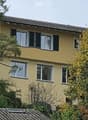 Two-story yellow house with multiple windows and a tiled roof, surrounded by trees and shrubs.