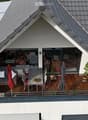 Wooden balcony with grey sofa, dining table, chairs, umbrella, and potted plants.