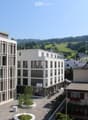 Modern multi-story building with large windows and balconies, surrounded by greenery and a scenic view of hills and trees.