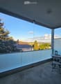 Balcony with glass railing, table, and chair, overlooking a scenic view of trees and a house.