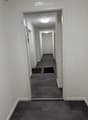 Empty hallway with white walls, gray carpeted floor, and recessed lighting. Two doors visible on the left side, with small rugs placed in front of them.
