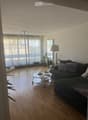 Modern living room with wooden flooring, large windows with sheer curtains, a dark grey sectional sofa with patterned cushions, a round glass coffee table, two floor lamps, and potted plants.