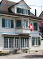 Two-story house with white walls, green shutters, and a red Swiss flag on the balcony. The house features a covered entrance with steps, potted plants, and a bench. The roof is tiled, and there are trees and shrubs in the surrounding garden area.