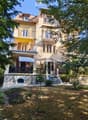 Two-story house with yellow and white exterior, multiple balconies, stone base, and lush greenery surrounding the property.