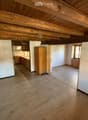 Empty room with wooden floor and exposed wooden ceiling beams. Adjacent kitchen area visible with cabinets and appliances.