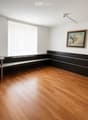 A modern, minimalist room with wooden flooring, a black chair, a small table, a mounted screen, and a framed picture on the wall.