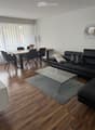Bright, modern living and dining area with hardwood flooring, large windows with sheer curtains, a black sectional sofa, a glass coffee table, a dining table with six chairs, a floor lamp, and a decorative picture frame on the wall.
