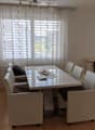 Bright living room with a sofa, coffee table, dining table, chairs, and a handbag on the floor. Large windows with white curtains and a modern painting on the wall.