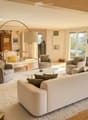 Modern living room with large windows offering a scenic view, featuring a cream-colored sofa, coffee table, armchairs, ottoman, floor lamp, dining table, and cabinet.