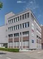 Modern multi-story building with large windows and brown accents, featuring signage for "Arbeitszentrum Liebfeld" and other companies. The building has a clean, contemporary design with a mix of light and dark tones.