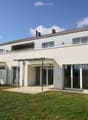 Modern two-story house with white exterior, large windows, and a balcony on the upper level. The house features a covered patio area with outdoor furniture and a well-maintained lawn.