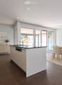 Modern kitchen and dining area with white cabinets, a central island, and a dining table with chairs. Large windows and sliding doors lead to a balcony.