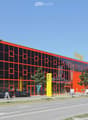 Modern, multi-story building with orange and black facade, large glass windows, and a parking area with several cars.