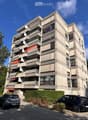 Modern multi-story apartment building with concrete facade, balconies, and large windows. Two cars are parked in front.