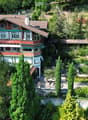 Aerial view of a large, multi-story house with red and white exterior, surrounded by lush greenery and trees. The house features multiple windows, a prominent roofline, and a well-maintained garden area with pathways and outdoor seating.