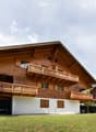 A large, multi-story wooden house with balconies and a spacious lawn in front, set in a scenic mountainous area.