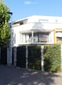 Modern two-story white house with a raised ground floor, featuring a balcony with outdoor furniture and an umbrella, surrounded by greenery and a hedge.