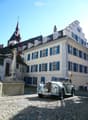 A large, multi-story building with white walls and blue shutters, featuring a classic car parked in front on a cobblestone street.