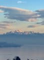 Panoramic view of a lake surrounded by mountains under a partly cloudy sky, with a few houses visible on the shore.