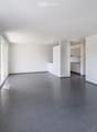 Empty room with white walls, dark tiled floor, large windows, and a small kitchen area with cabinets and appliances visible in the background.