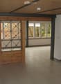 Empty room with tiled floor, white walls, wooden ceiling beams, and large windows with metal-framed glass panels.