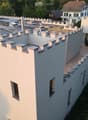 Modern two-story building with a distinctive, castle-like design featuring crenellated roofline and multiple chimneys. The structure includes a wooden deck area, glass railing, and surrounding greenery.