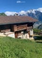 A wooden chalet with a brown tiled roof, wooden balcony, and stone base, situated on a grassy slope with a stunning mountain backdrop.