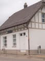 Two-story building with a tiled roof, white walls, and multiple windows. Features a sign reading "Gontenschwil" and a small entrance area with steps.