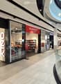 Shopping mall interior with glass storefronts, clothing racks, shelves, and a cashier counter visible inside a store.